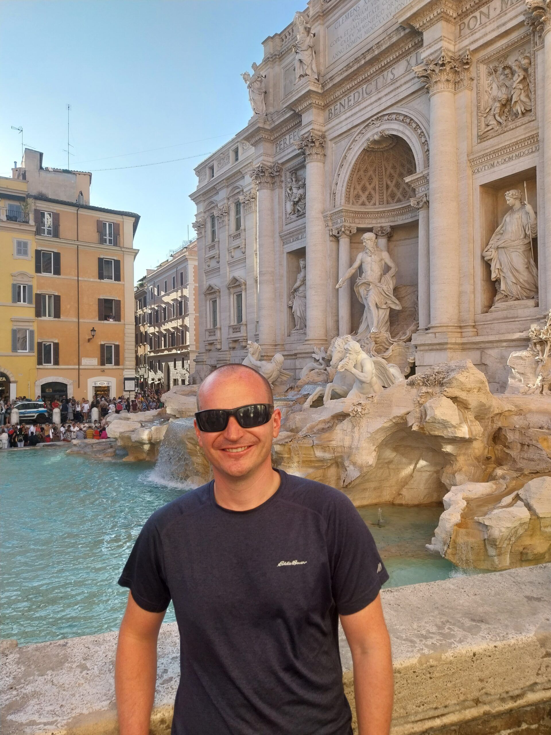 Man in a dark t-shirt and sunglasses stands smiling in front of the Trevi Fountain in Rome, with ornate statues behind him.