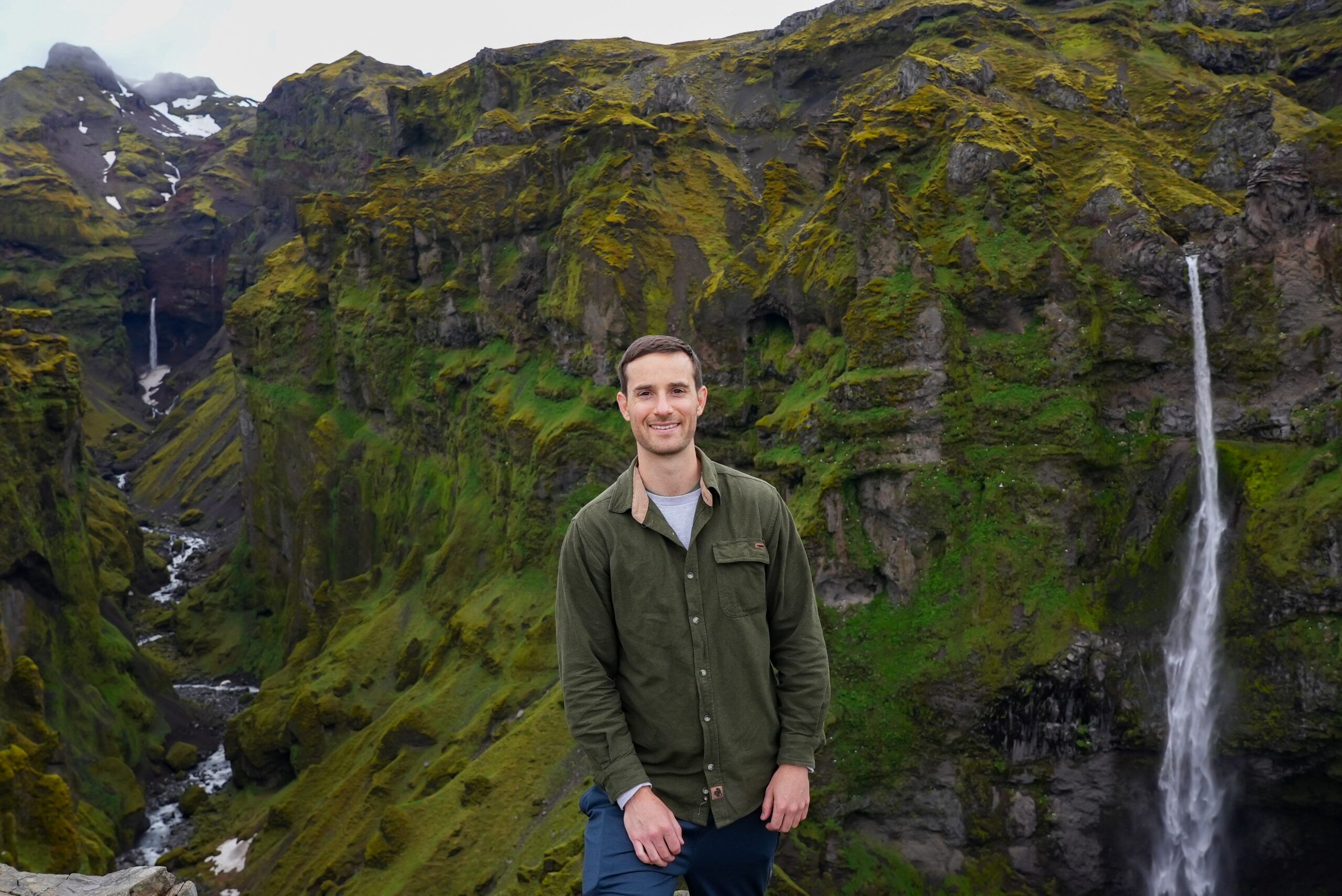 Man smiling in front of a mossy canyon with a tall waterfall on the right background, outdoors.