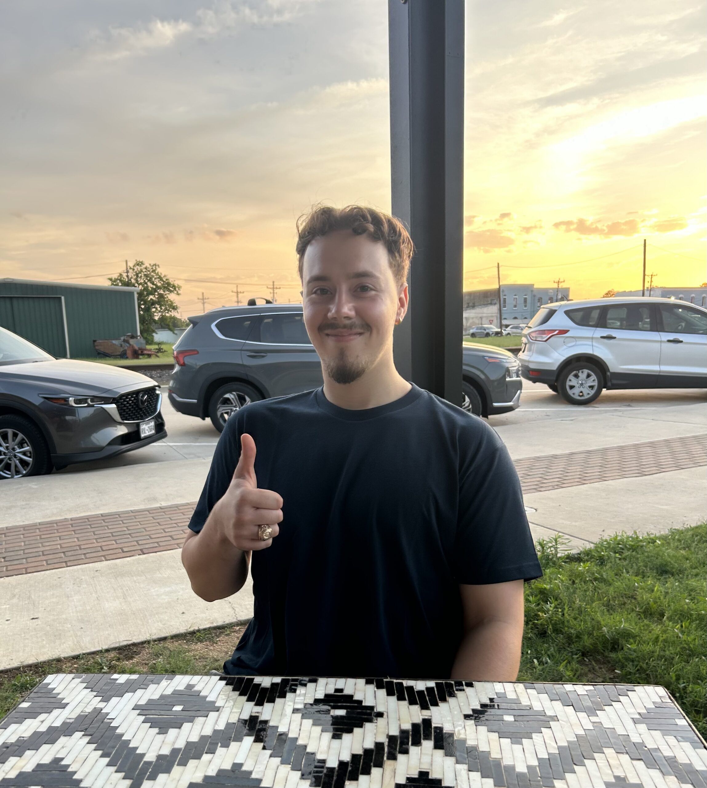 Man with short hair and goatee smiles and gives a thumbs up while seated at a mosaic table outdoors at sunset, wearing a black shirt.