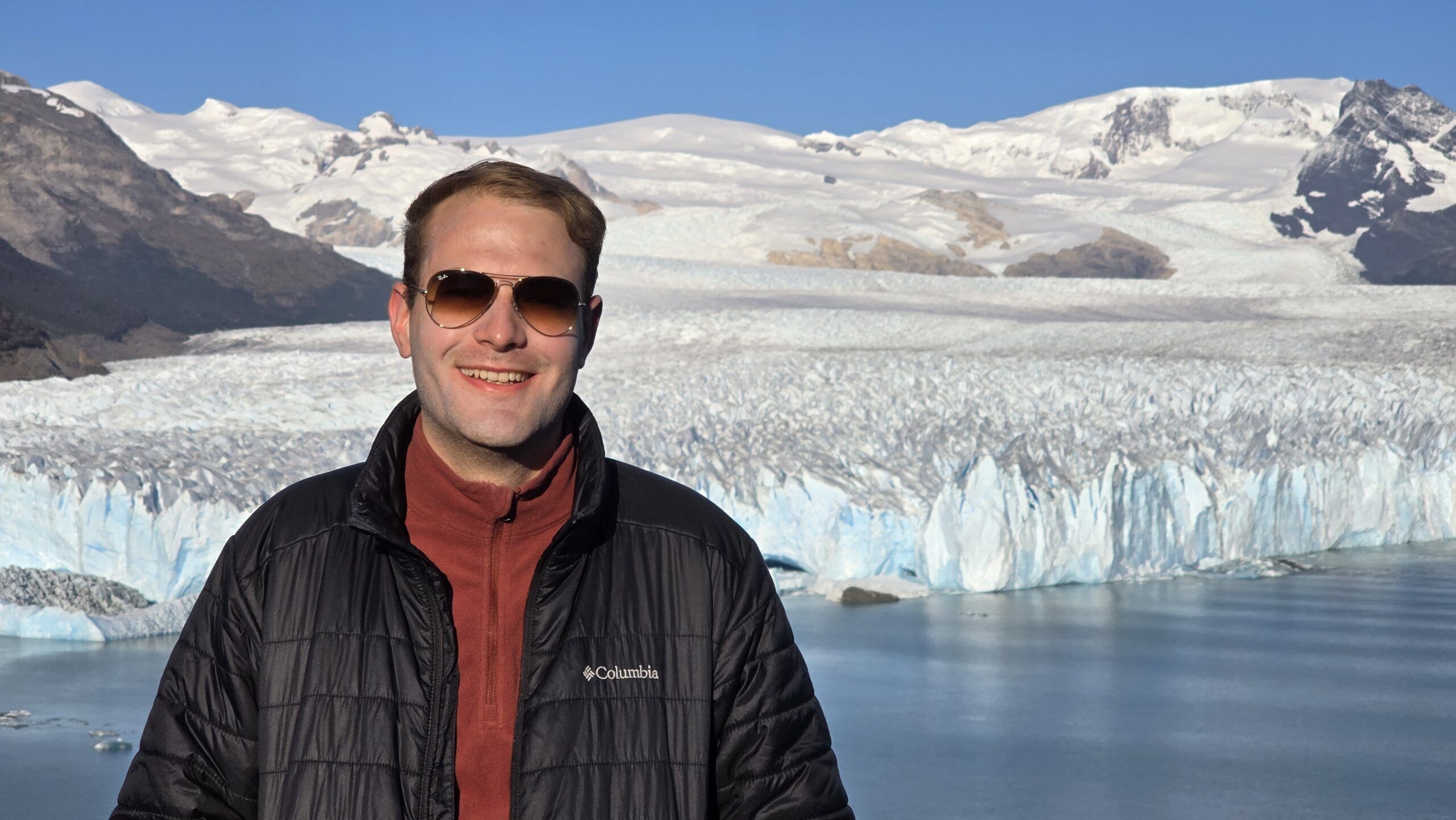 Man in sunglasses and a black jacket posing in front of a snowy glacier and blue sky.