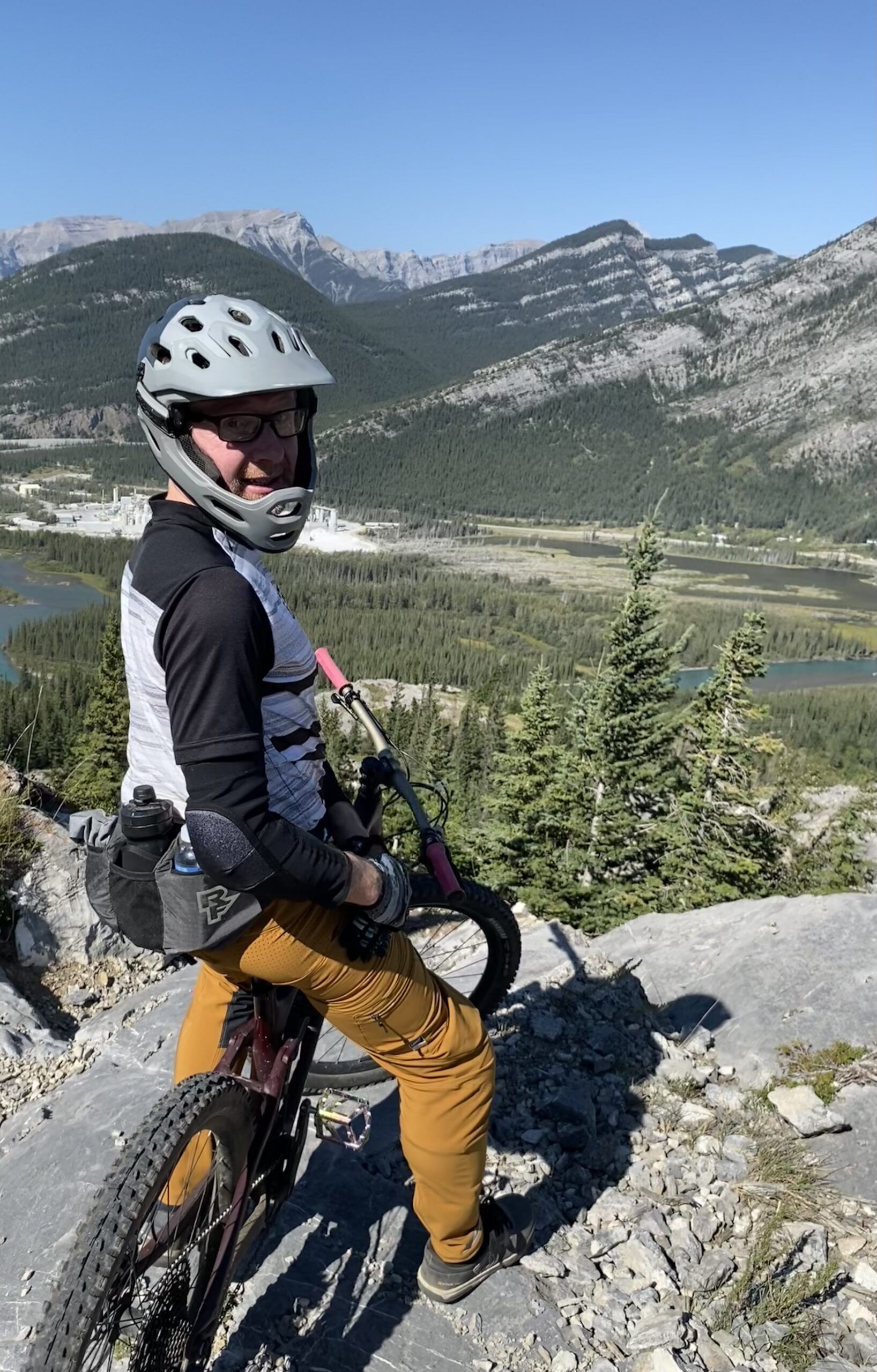 Mountain biker wearing a white helmet and protective gear pauses on a rocky trail with a forested valley and mountains in the background.