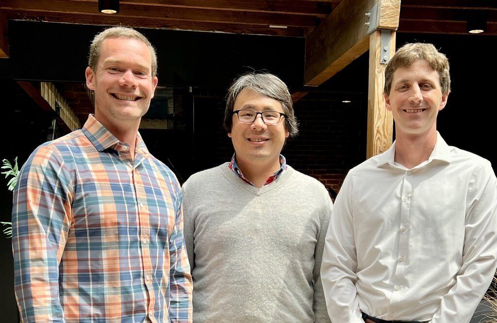Three men standing side by side indoors, smiling, in a casual office-like space with wood beams above them.